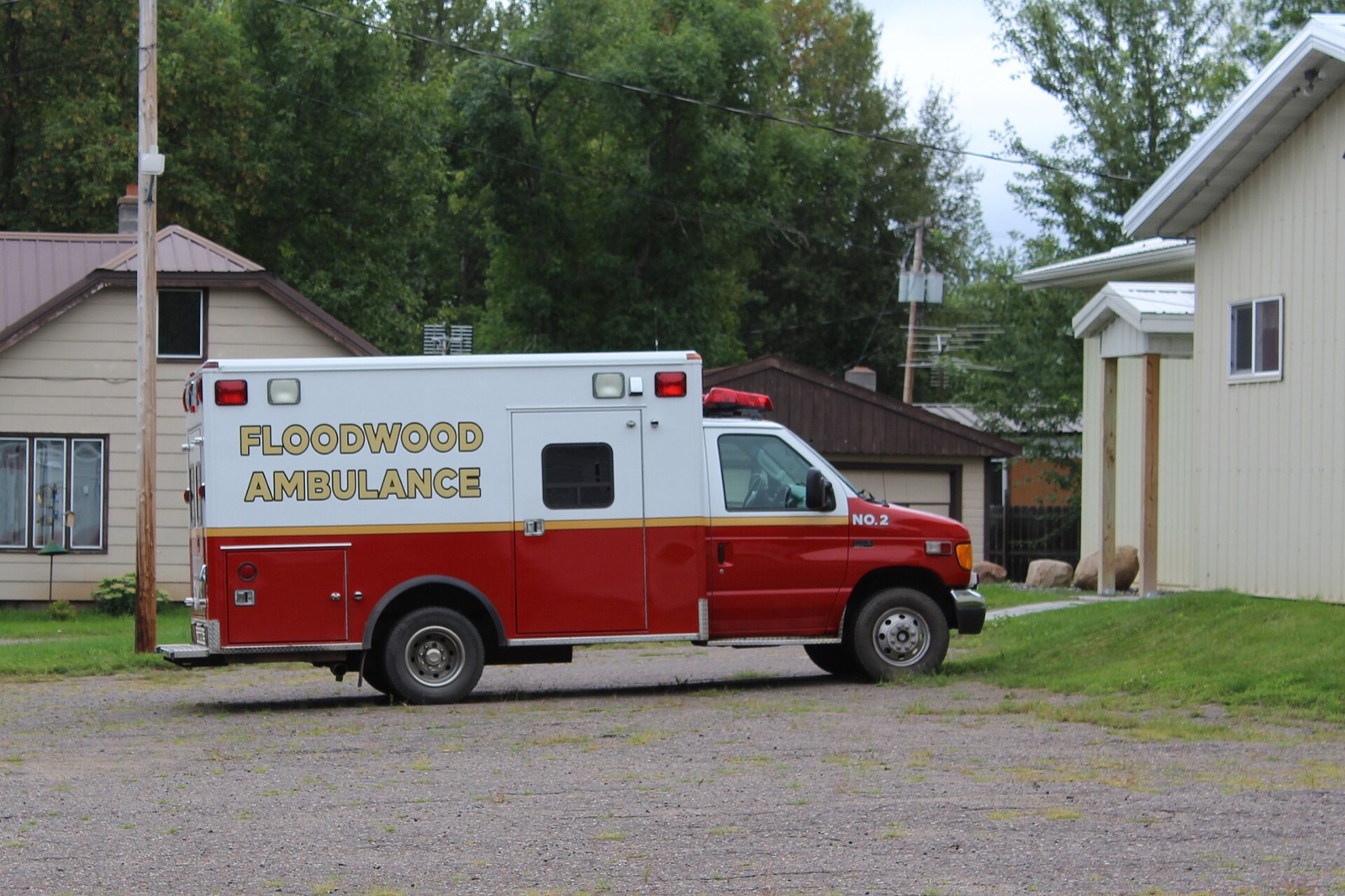 A red and white ambulance marked Floodwood Ambulance, No. 2, parked on a gravel lot beside small town buildings.